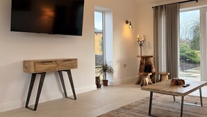 Ein modernes Wohnzimmer mit Flachbildfernseher an der Wand, einem Holzkonsolentisch, einem Couchtisch und einem großen Fenster mit Vorhängen.