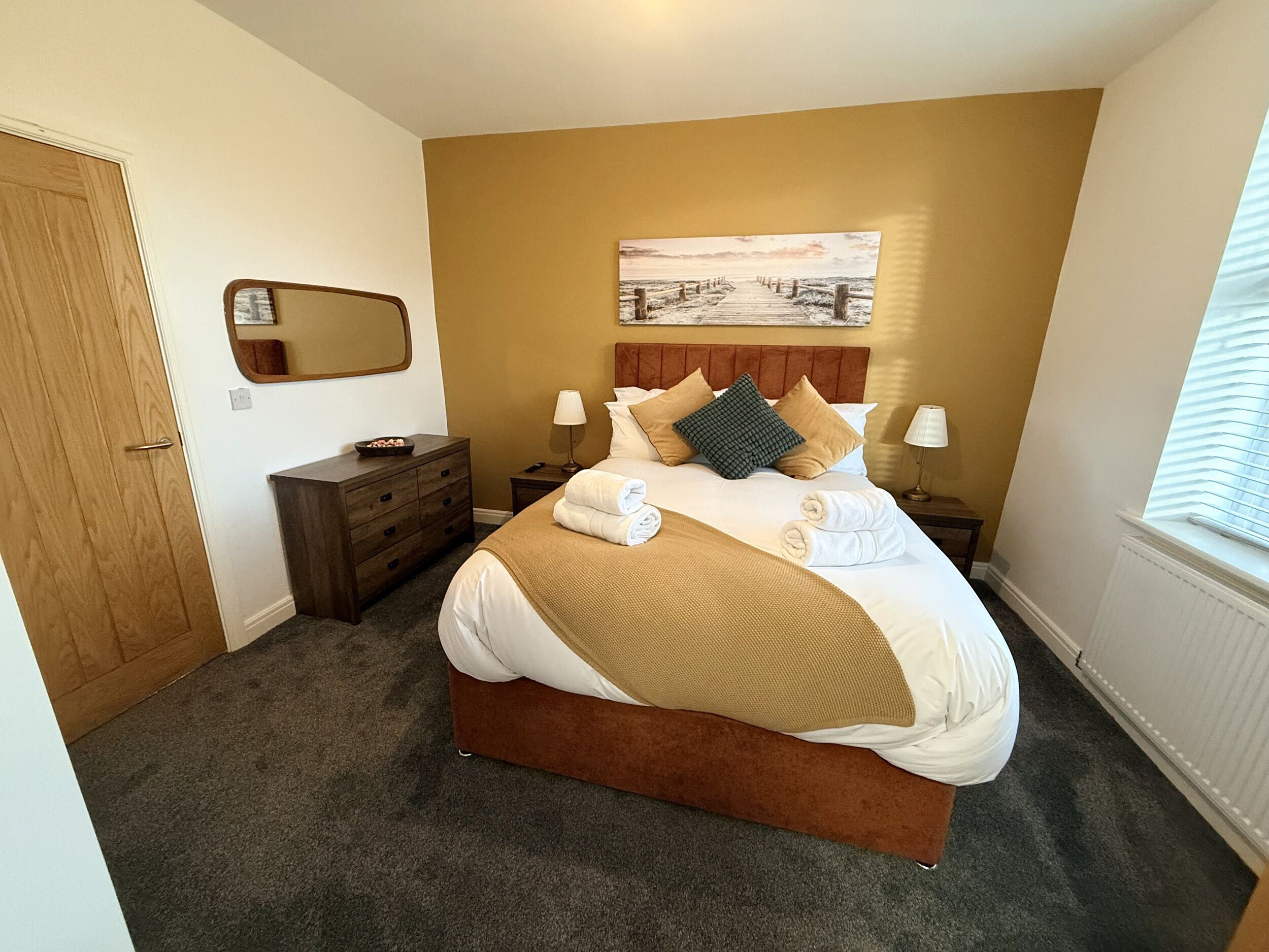 A bedroom with a large bed, two bedside tables, a nightstand, a wooden dresser, a mirror, and a window with blinds.