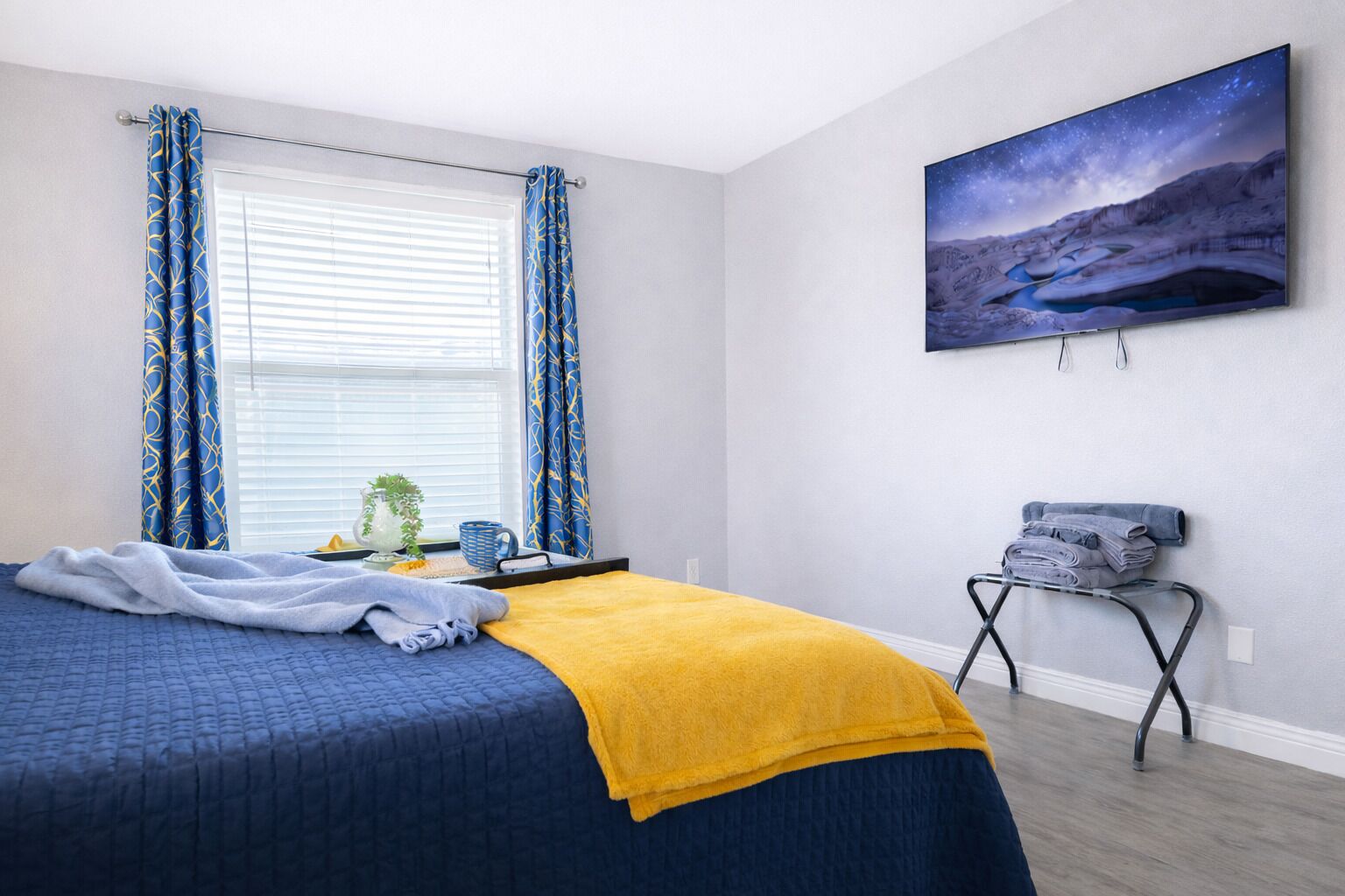 Une chambre à coucher comprenant un lit, une table de chevet, une fenêtre avec des rideaux, un téléviseur fixé au mur et une petite planche à repasser.