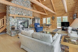 Ein gemütliches Blockhaus-Interieur mit Kamin aus Stein, Holzbalken, einer Couch, einem blauen Sessel und einem Fernseher.