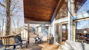 Une terrasse en bois avec des fauteuils Adirondack et un banc, un espace couvert doté d’un plafond en bois, et une porte vitrée donnant sur une pièce contenant une table et des chaises.