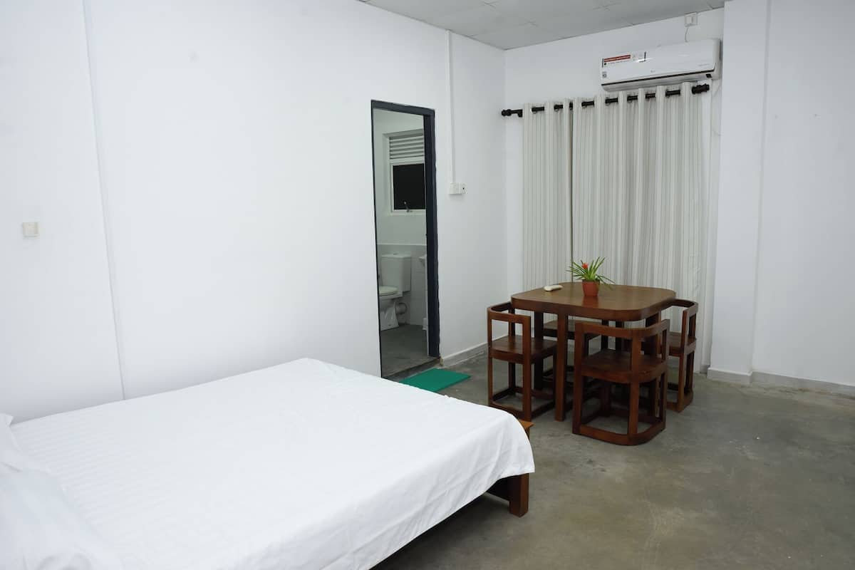Una habitación pequeña y limpia con una cama individual, una mesa de madera con sillas, una planta en maceta y una ventana con cortinas.
