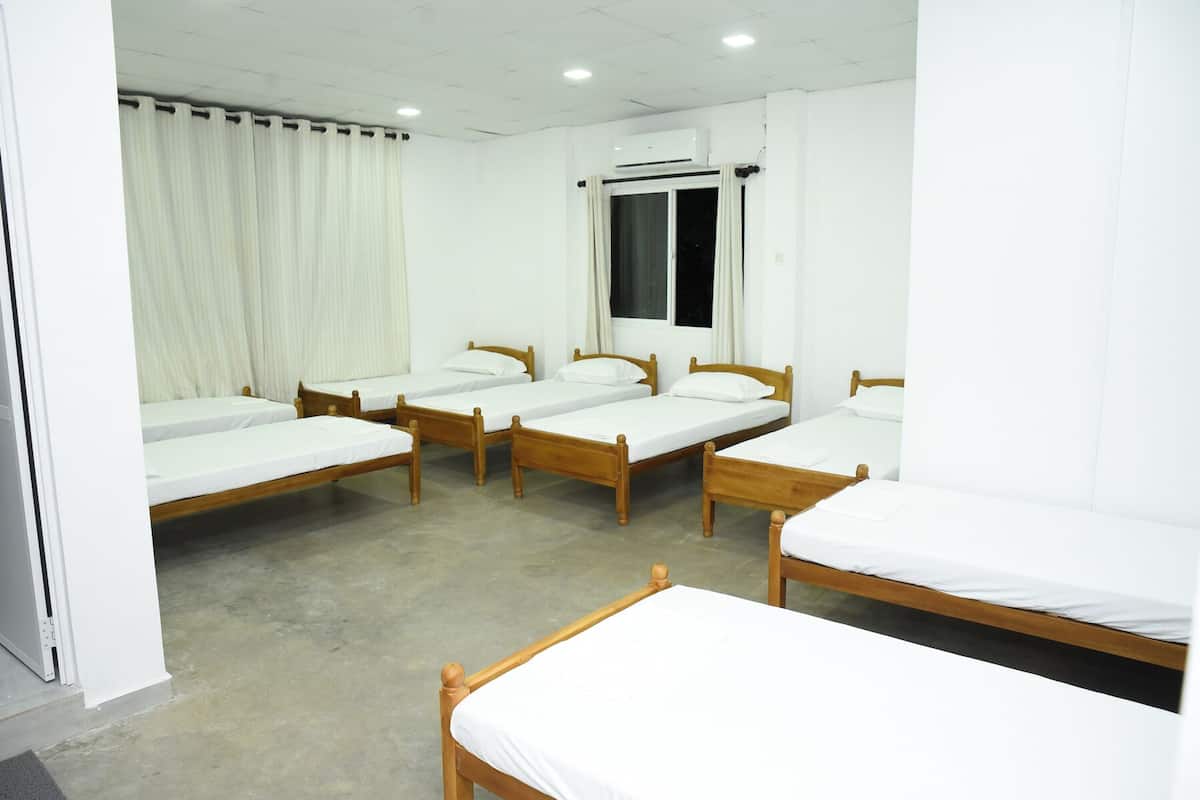 Habitación de hotel con cuatro camas de madera, ropa de cama blanca, una ventana con cortinas, equipo de aire acondicionado y piso de concreto.