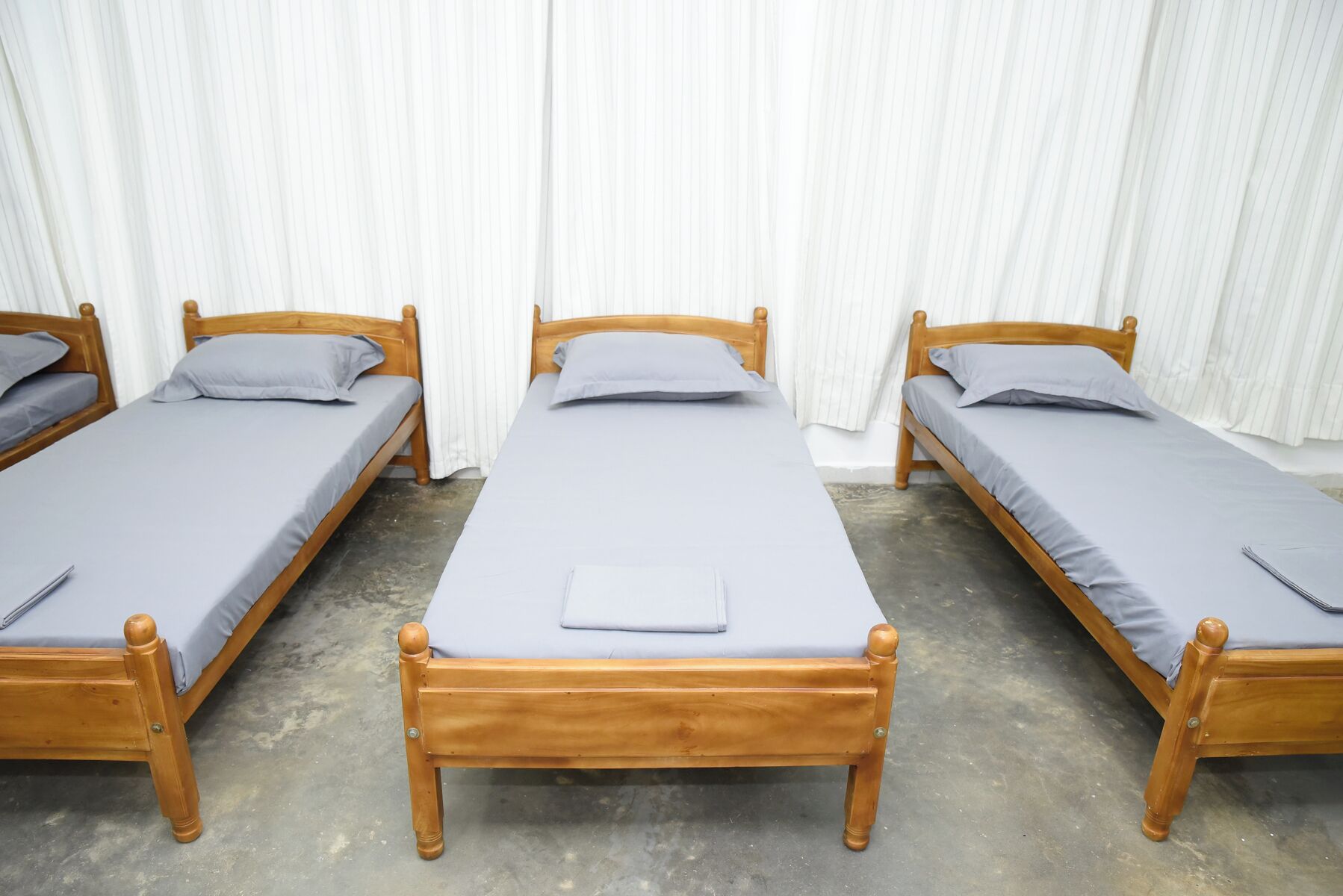 A row of three wooden single beds with grey bedding, arranged in a row against a white curtain backdrop.