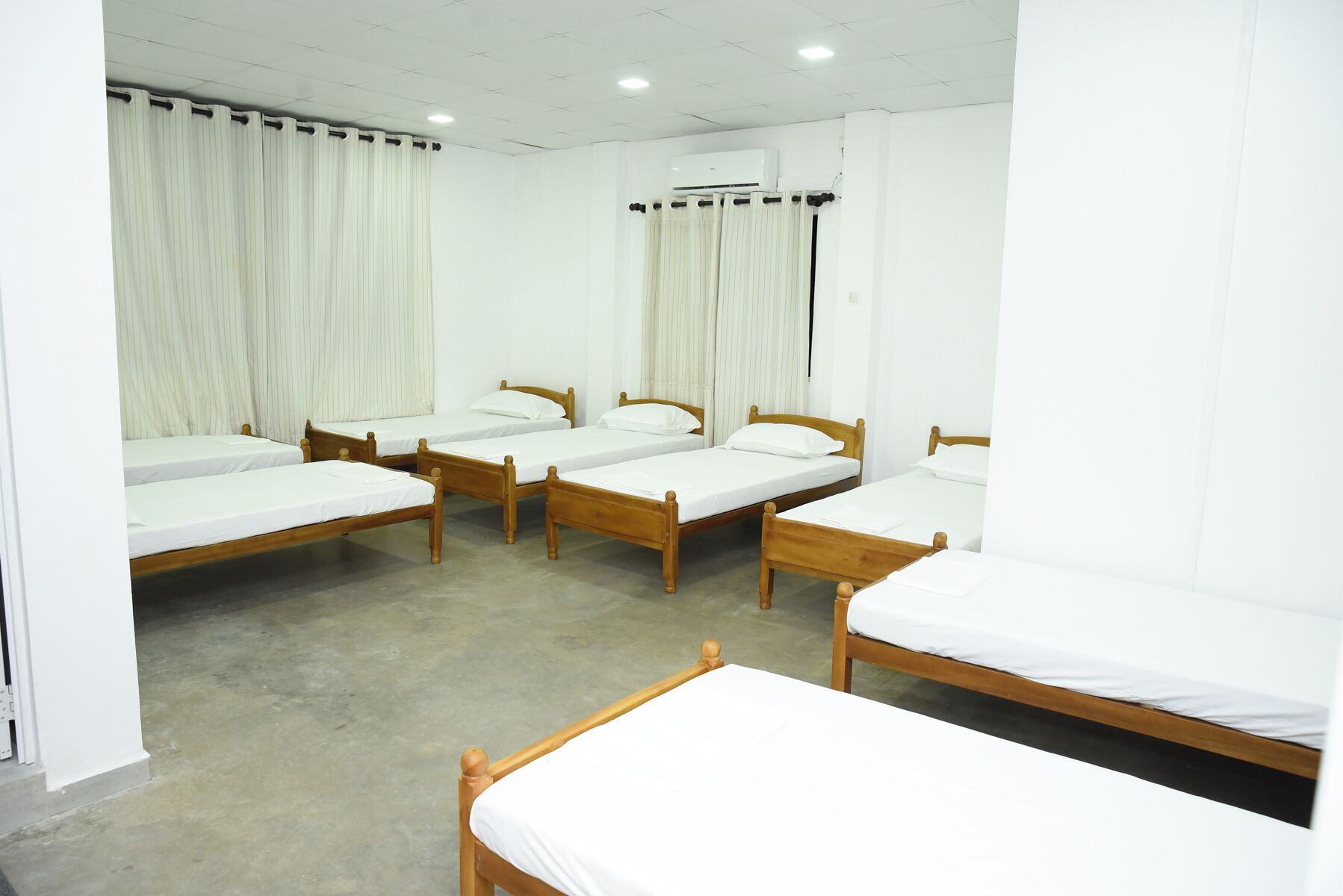 A room with four wooden beds, white curtains, and an air conditioning unit on the wall.