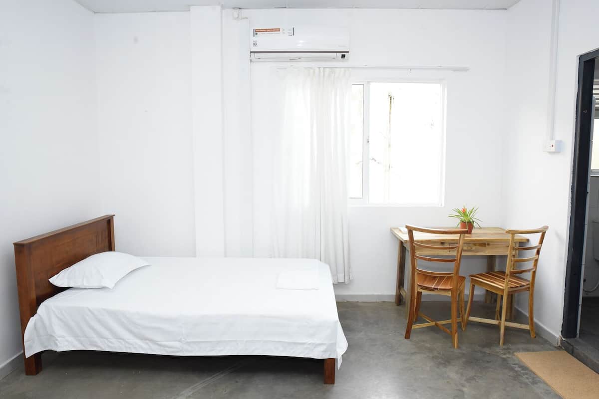 Un dormitorio con una cama de madera, una pequeña mesa de madera con sillas, una ventana con cortinas y un equipo de aire acondicionado.