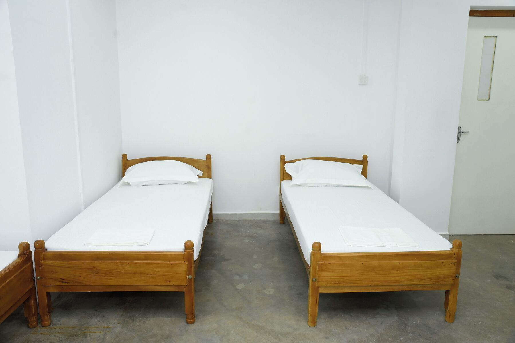Two wooden single beds with white bedding in a room with plain white walls and a concrete floor.