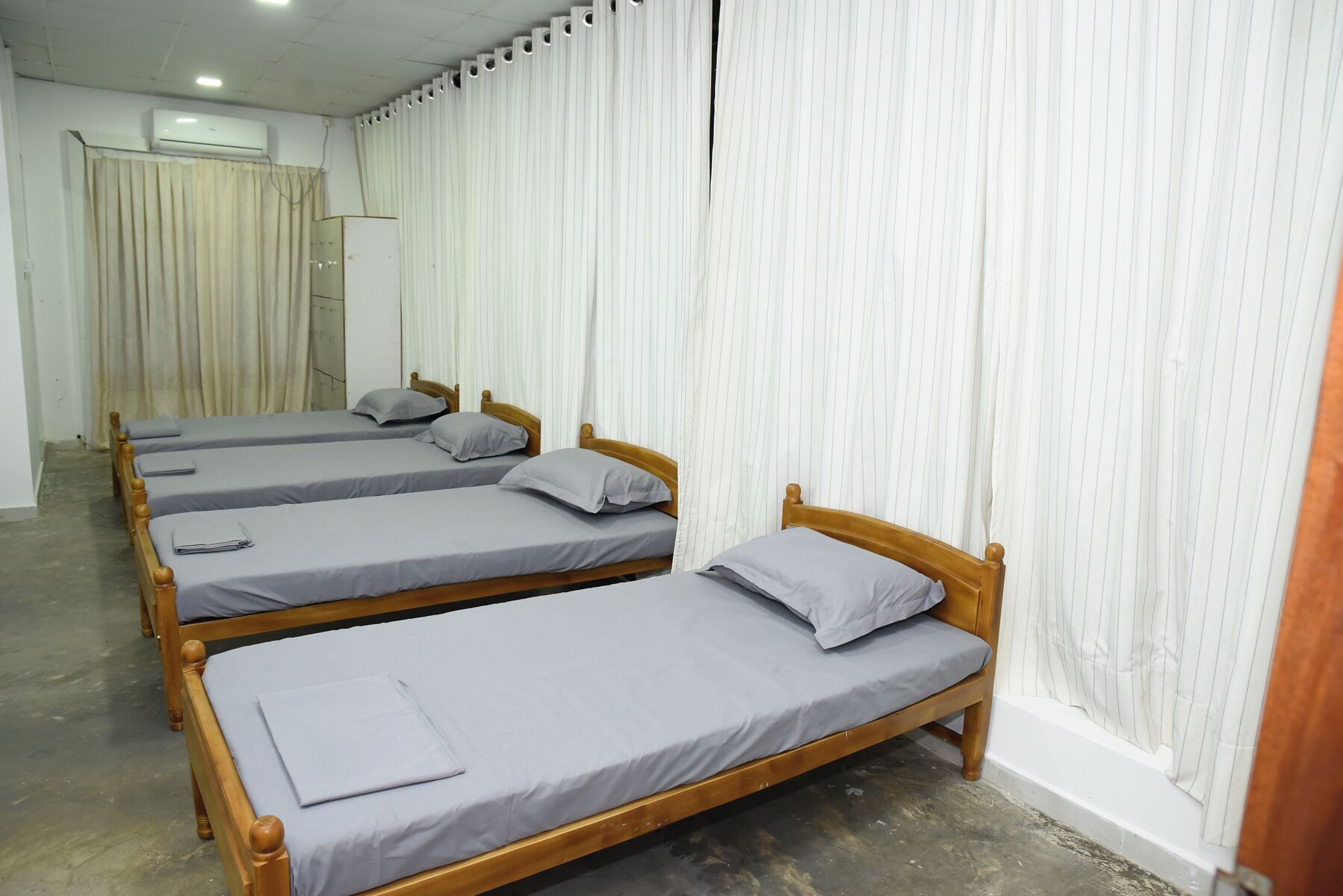 A room with four wooden beds aligned against a white curtain wall, each with a grey mattress and pillow.