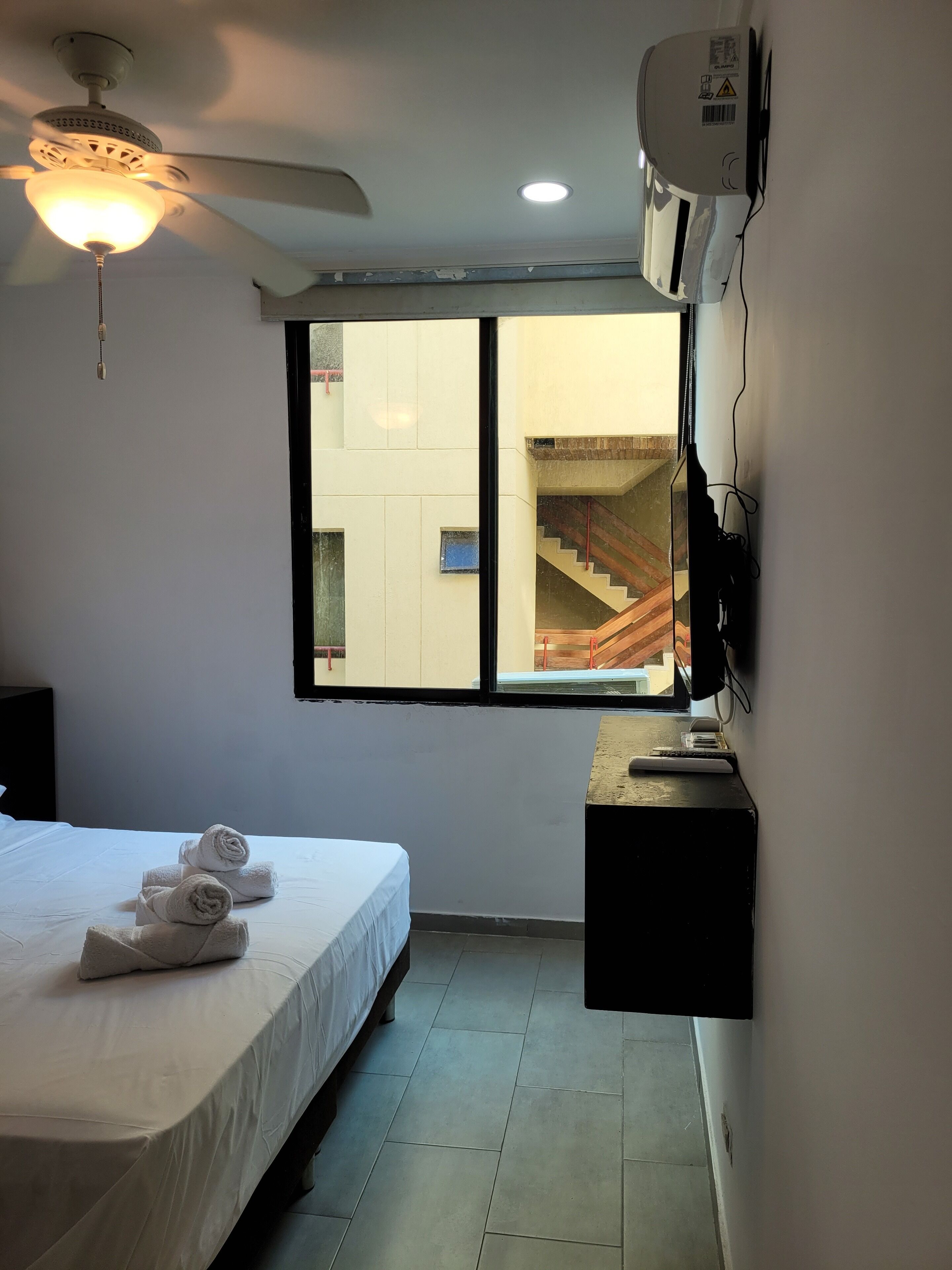 A hotel room with a bed, a ceiling fan, a window with a view of a staircase, and an air conditioning unit.