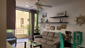 Un soggiorno con divano, tavolino in vetro, ventilatore a soffitto, tavolo da pranzo con sedie e varie decorazioni a parete.