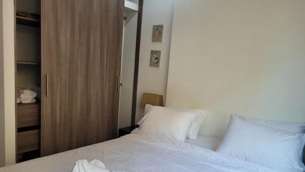 Una camera d'hotel con un letto, lenzuola bianche, un armadio in legno e un comodino con una lampada.