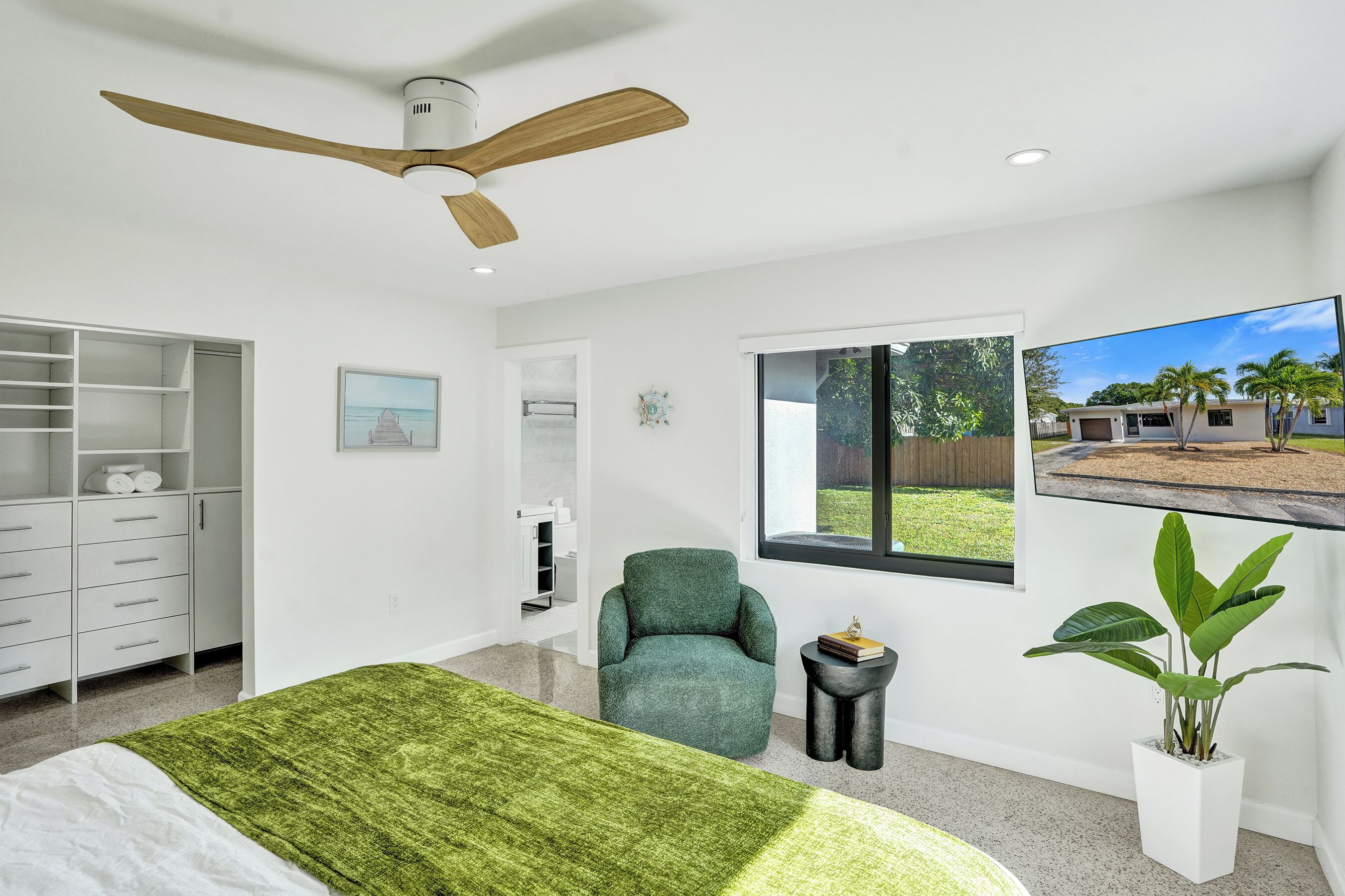 A modern bedroom with a bed, a green armchair, a ceiling fan, a window with a view of a house, and a potted plant.