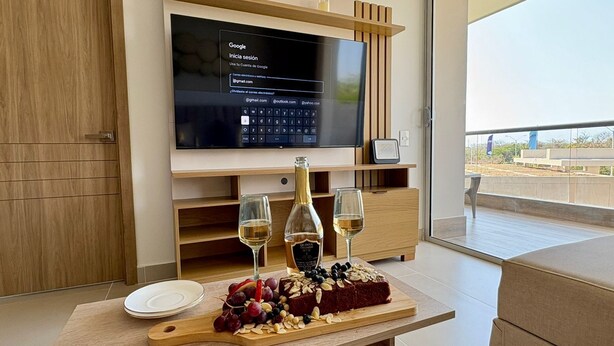 Een moderne woonkamer met een flatscreen televisie aan een houten muur, een houten eettafel met een schaal fruit en wijnkaraffen, en uitzicht op de omgeving door grote ramen.