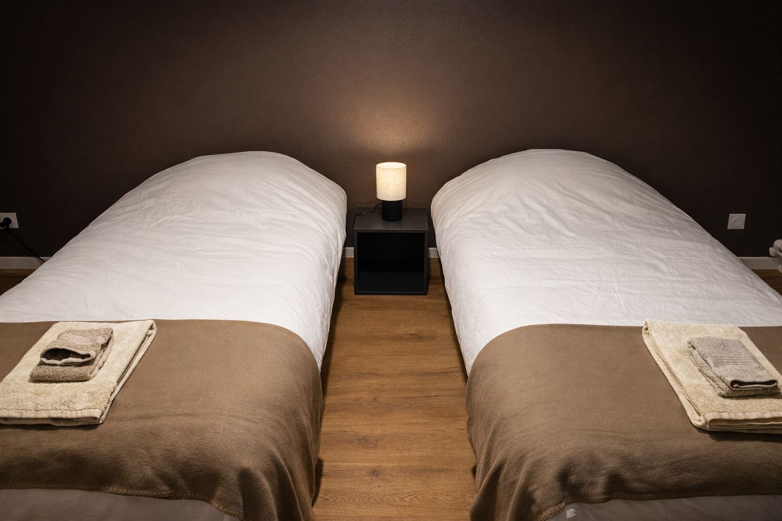 Una habitación con dos camas, una mesita de noche con lámpara y suelo de madera.
