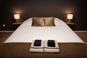 Ein ordentlich bezogenes Bett mit zwei Kissen, ein Paar gefaltete Handtücher und je eine Nachttischlampe auf Holzbeistelltischen.