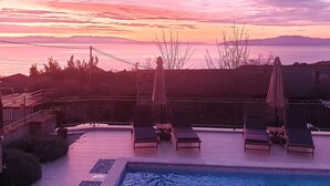 Ein Poolbereich mit Liegestühlen und Sonnenschirmen, umgeben von einem Zaun und Bäumen, mit Blick auf Berge im Hintergrund.