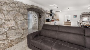 Ein modernes Wohnzimmer mit Steinwand, einer Couch und einer Küchenzeile mit weißen Schränken sowie einem Esstisch.