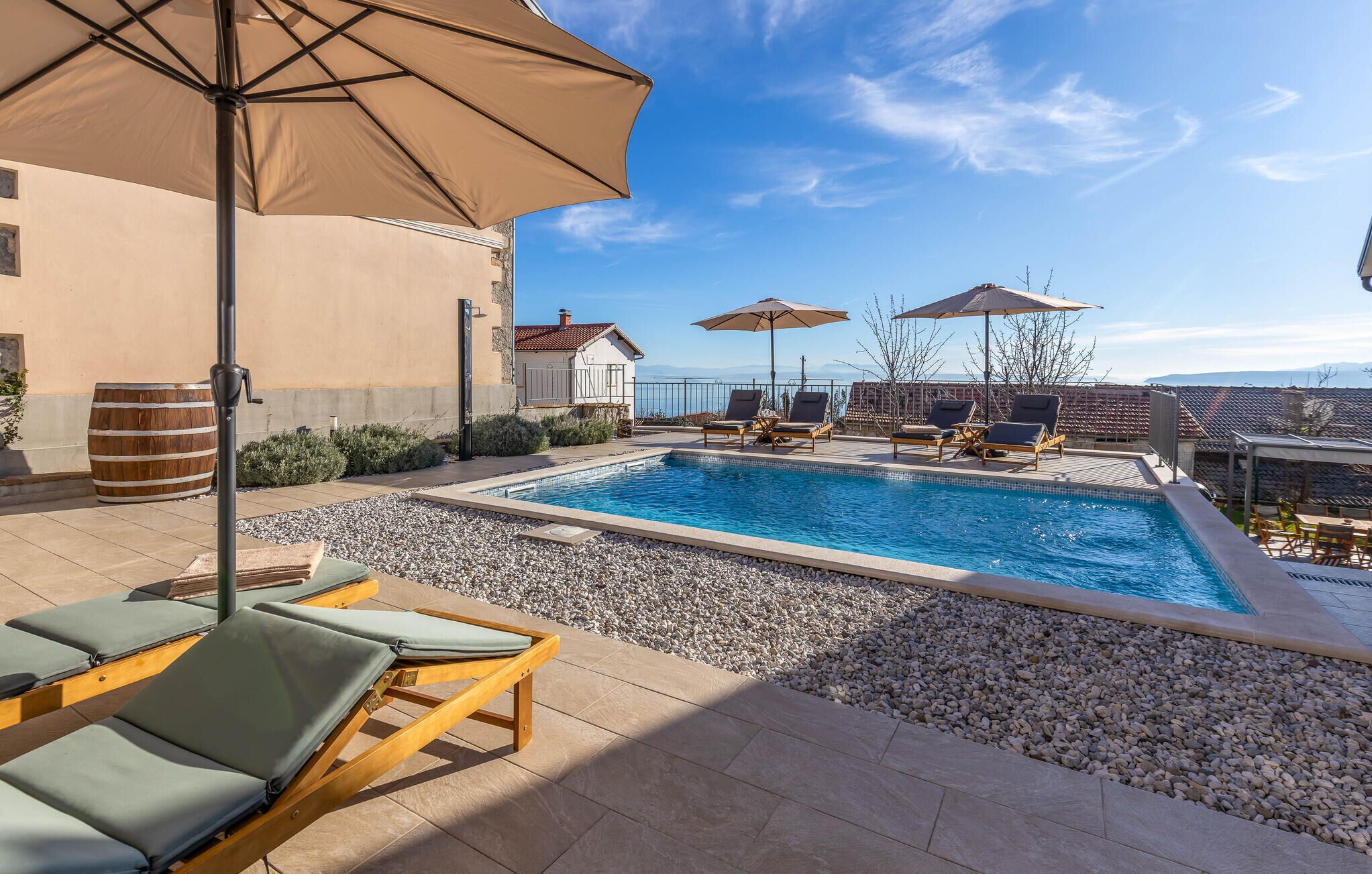A pool area with lounge chairs and umbrellas, surrounded by a pebble border and a building with a tiled roof.