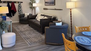A living room with a sofa, a blue armchair, a dining table with chairs, a lamp, a TV, and a fireplace. - The Beckman Bungalow - Lakeside 2-bedroom Home (Delavan)