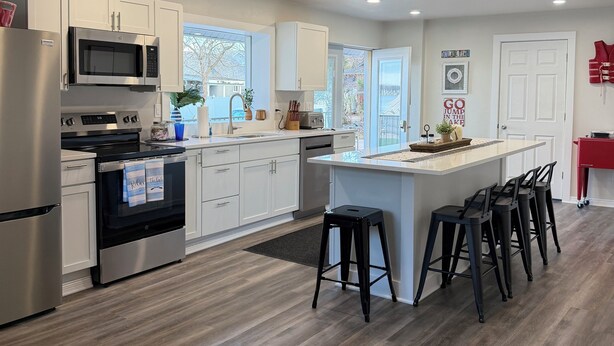 A modern kitchen with stainless steel appliances, a central island with bar stools, and a view of the outdoors. - The Beckman Bungalow - Lakeside 2-bedroom Home (Delavan)