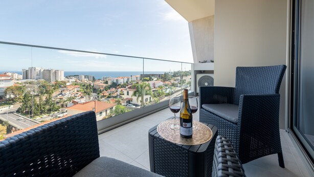 Un balcón con mobiliario de mimbre, una botella de vino y copas sobre una mesa, con vistas a un paisaje urbano.