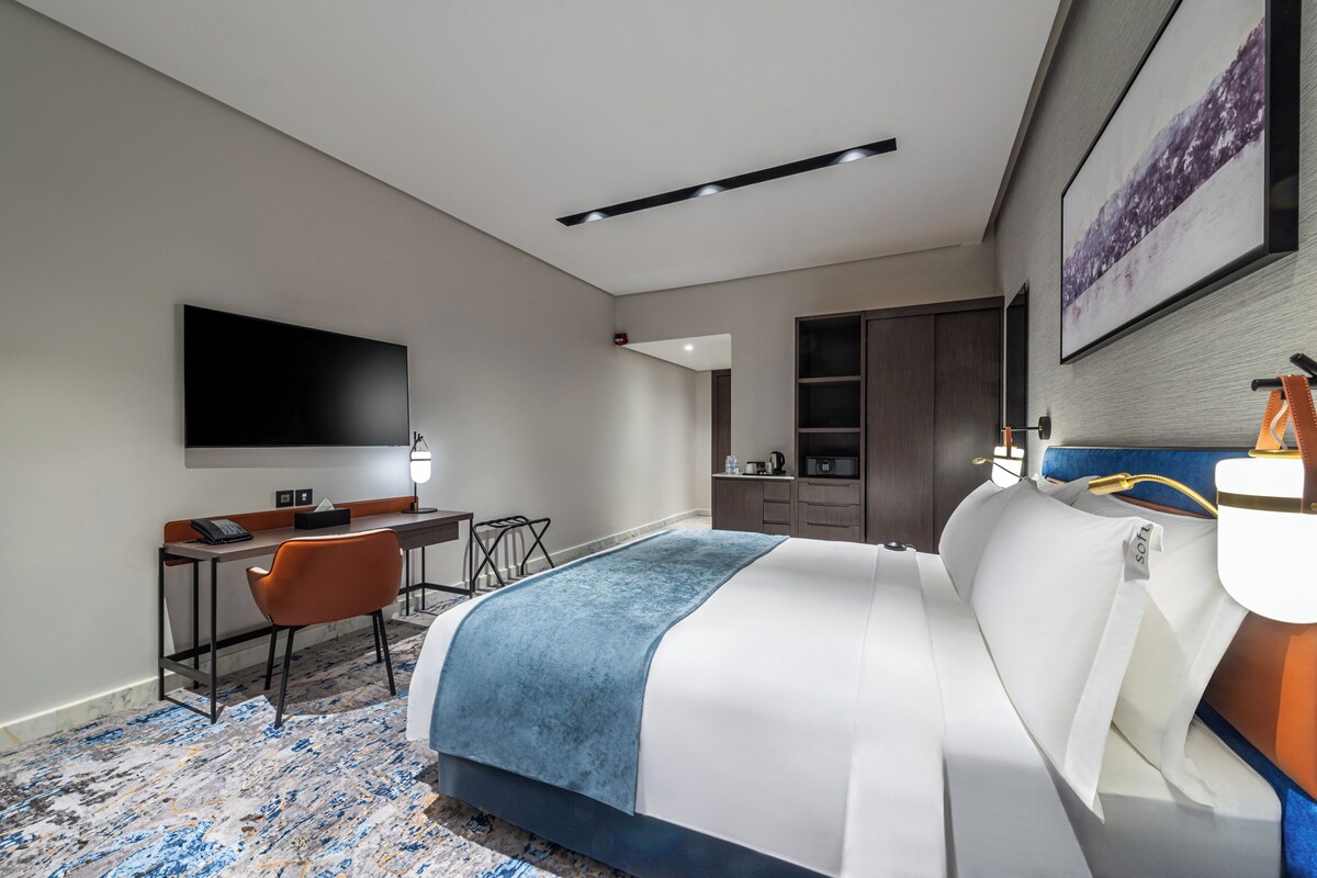 A modern hotel room with a large bed, a desk with a chair, a flat-screen TV, and a wardrobe.