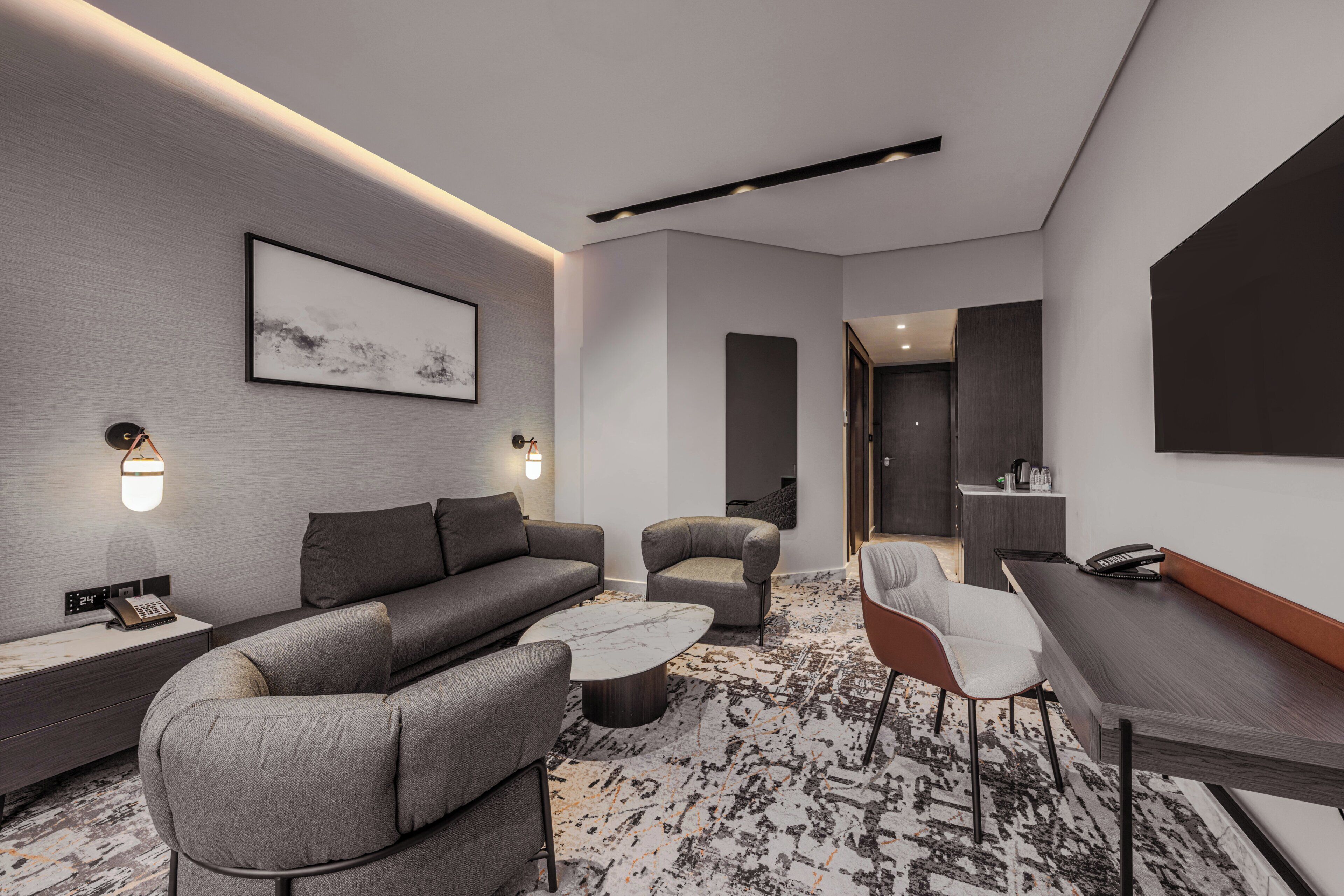 A modern hotel room with a sofa, armchair, coffee table, and a desk with a telephone.