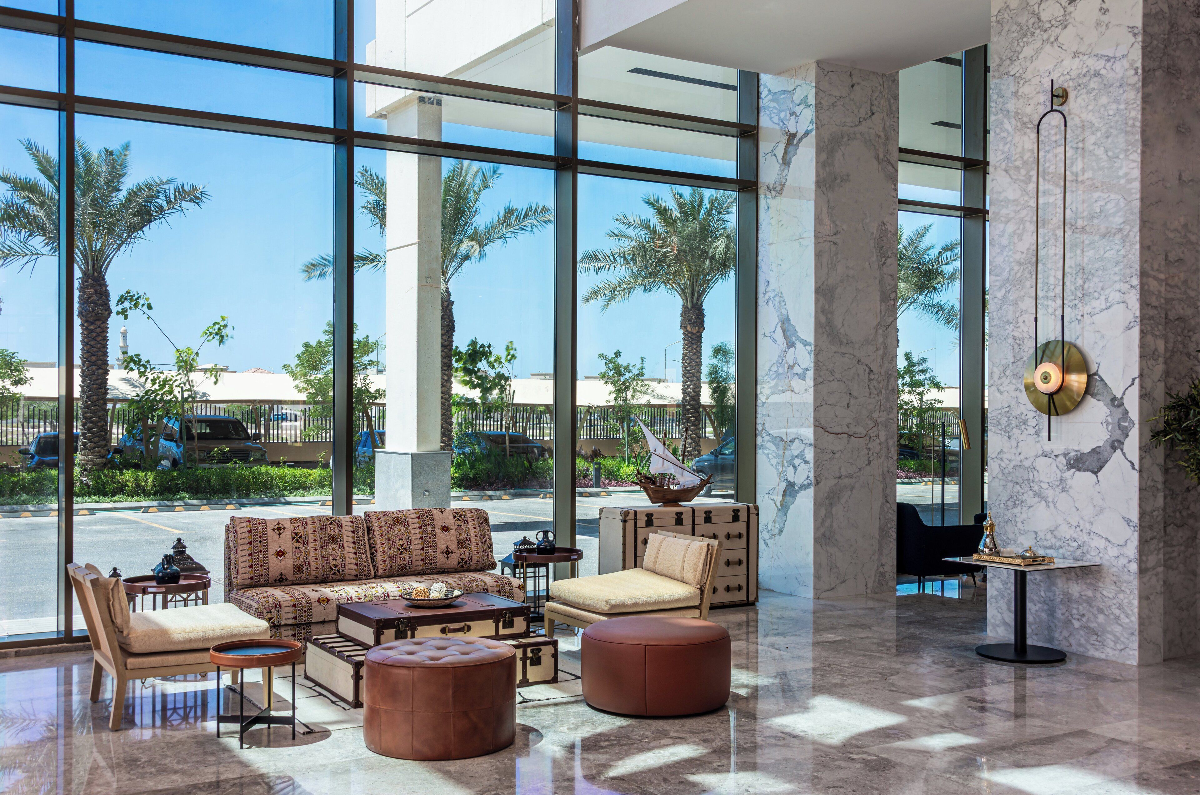 A spacious lobby with a large window offering a view of palm trees and a parking area. There is a seating area with sofas and ottomans, and a small table with decorative items.