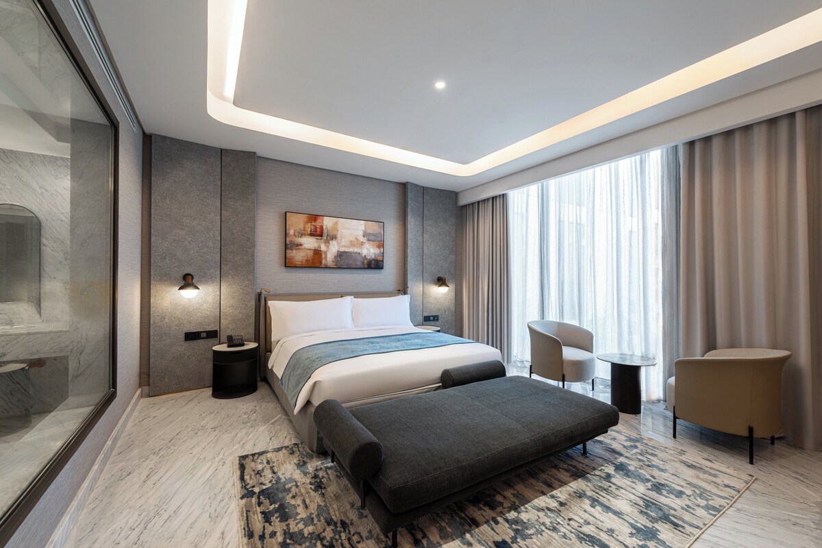 A modern hotel room with a large bed, a seating area with two chairs, a small table, and a large window with curtains.