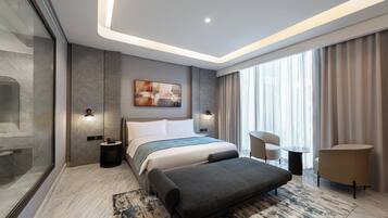 Quarto de hotel moderno com uma cama grande, área de estar com duas cadeiras, uma mesinha e uma janela ampla com cortinas.