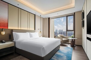 A modern hotel room with a large bed, a desk, a chair, a TV, and a view of the city.