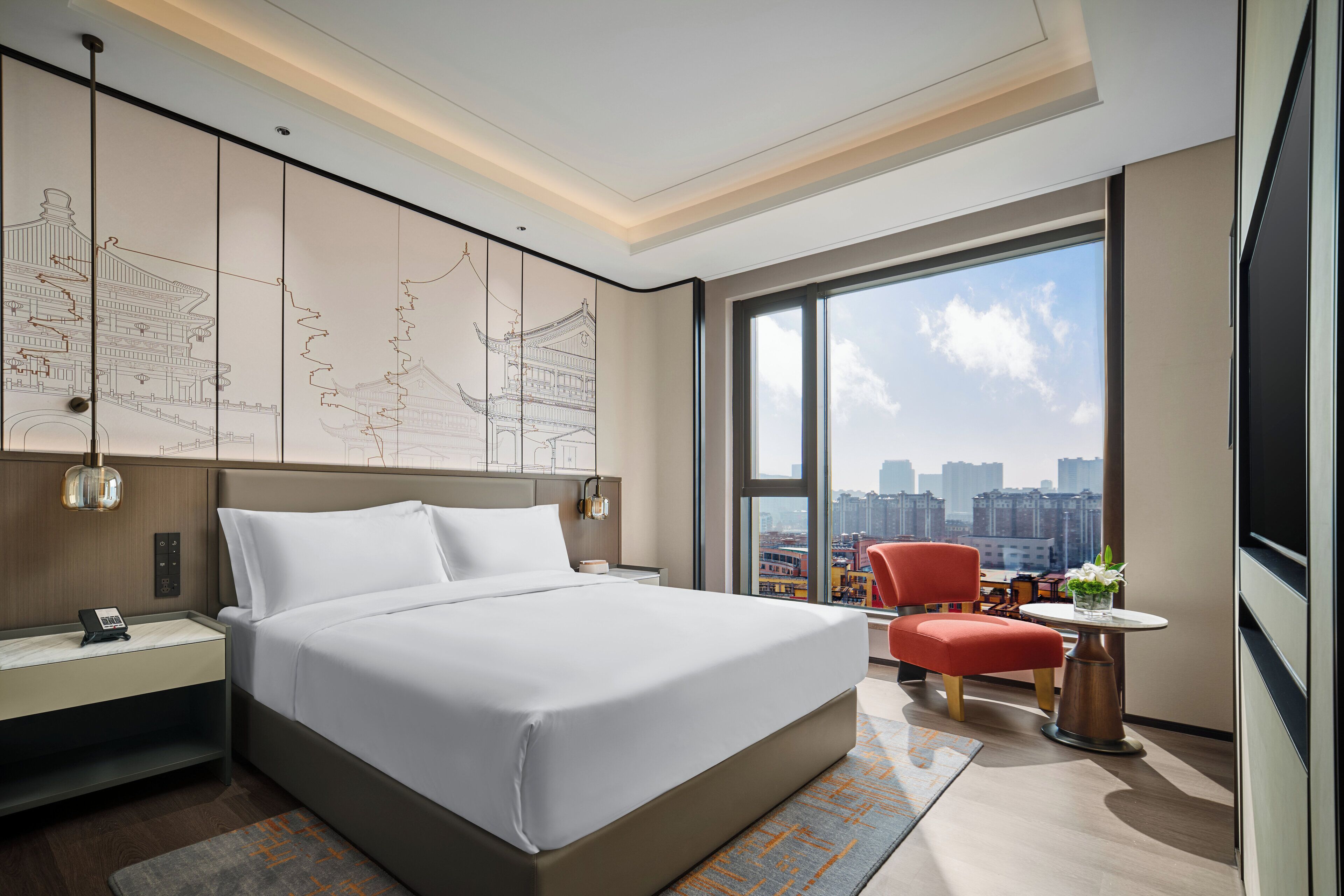 Une chambre d’hôtel moderne dotée d’un grand lit, d’un bureau avec un téléphone, d’une chaise, d’une petite table avec un vase et d’une grande fenêtre donnant sur la ville.