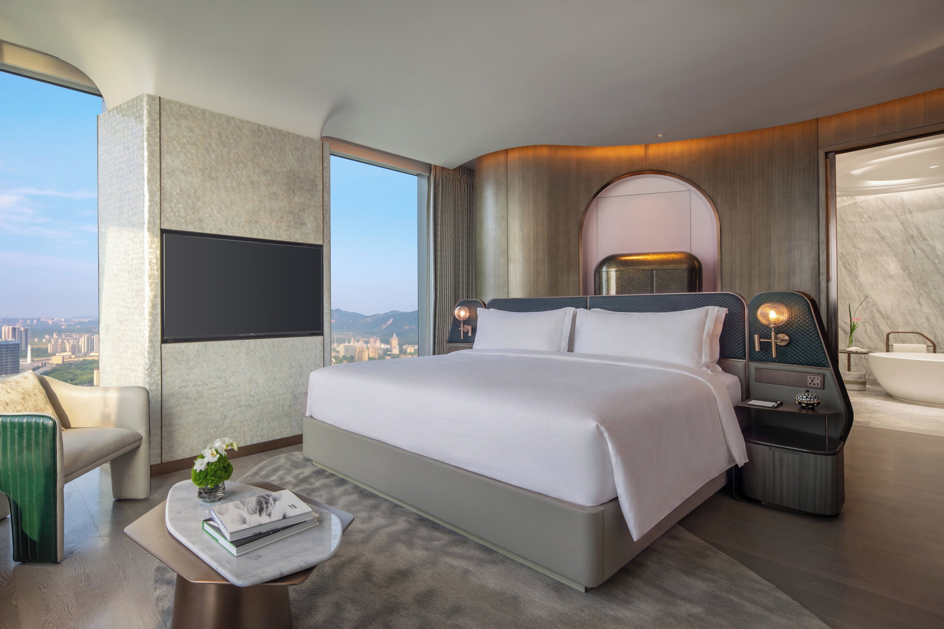 A modern hotel room with a large bed, a sitting area with a chair, a small table with a book and flowers, a nightstand with a lamp, and a bathtub.
