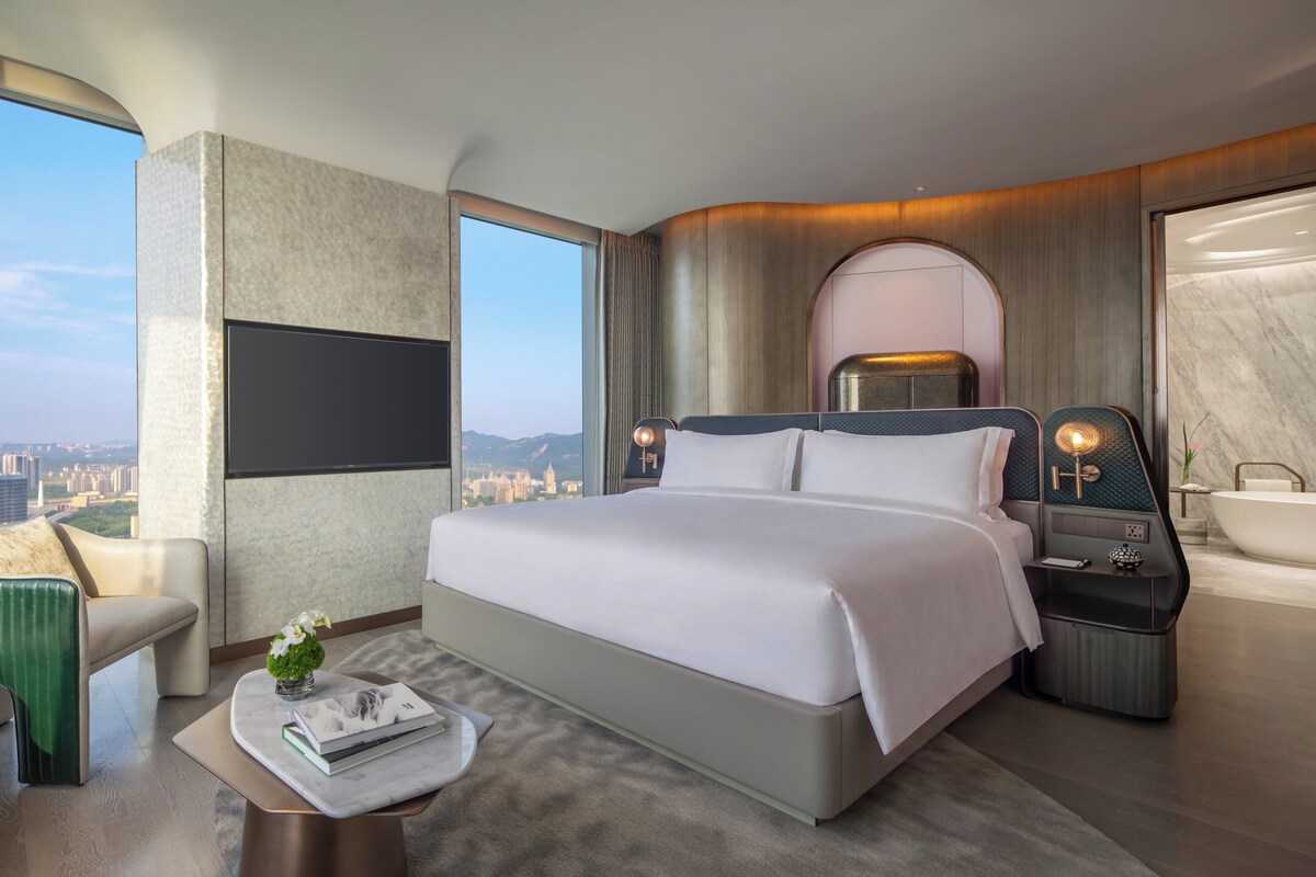 A modern hotel room with a large bed, a sitting area with a chair, a small table with a book and flowers, a nightstand with a lamp, and a bathtub.