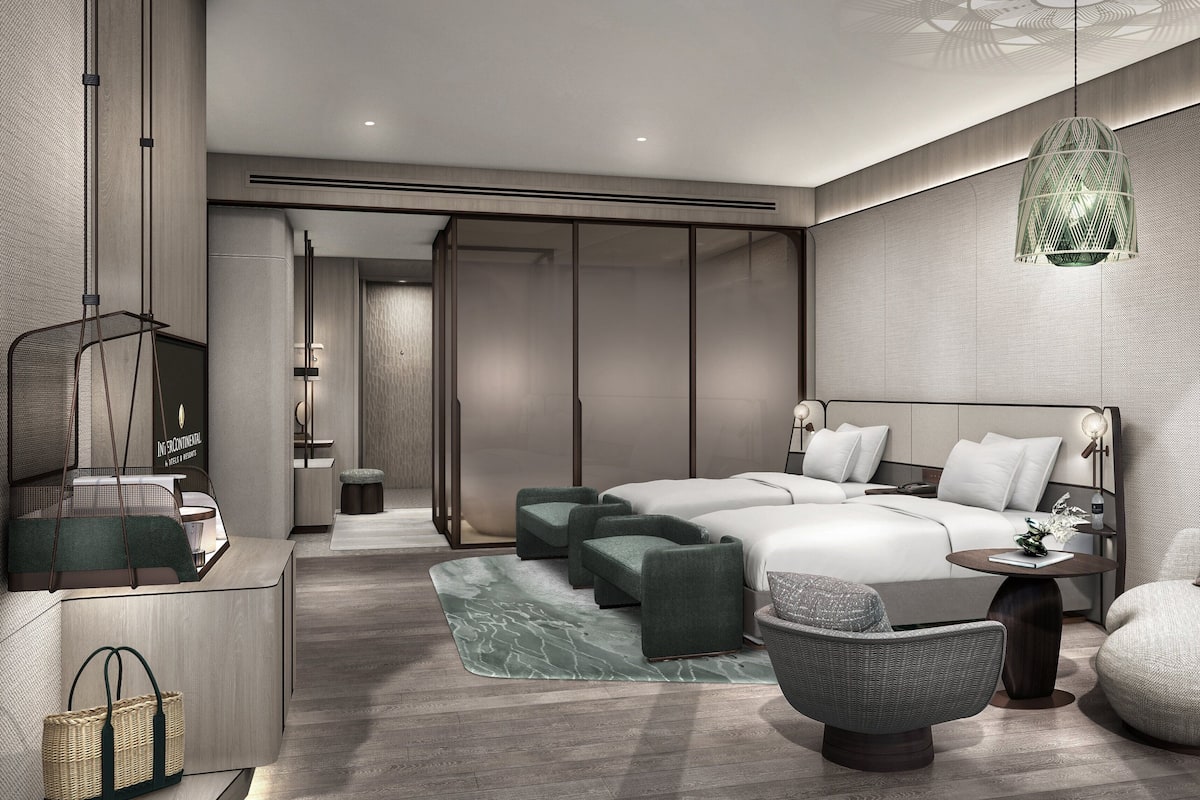 Ein modernes Hotelzimmer mit einem großen Bett, zwei Nachttischen, einer Pendelleuchte und einer Sitzecke mit Sessel und Hocker.