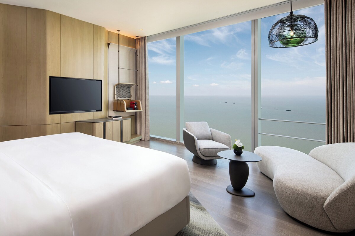 A hotel room with a large bed, a desk with a flat-screen TV, a chair, a small table with a vase, and a balcony with a view of the sea.