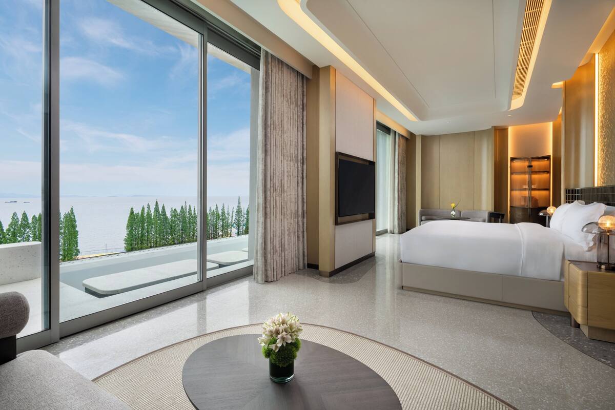 Ein modernes Hotelzimmer mit großem Fenster, einem Bett, einem Fernseher und einem kleinen Tisch mit Blumenvase.