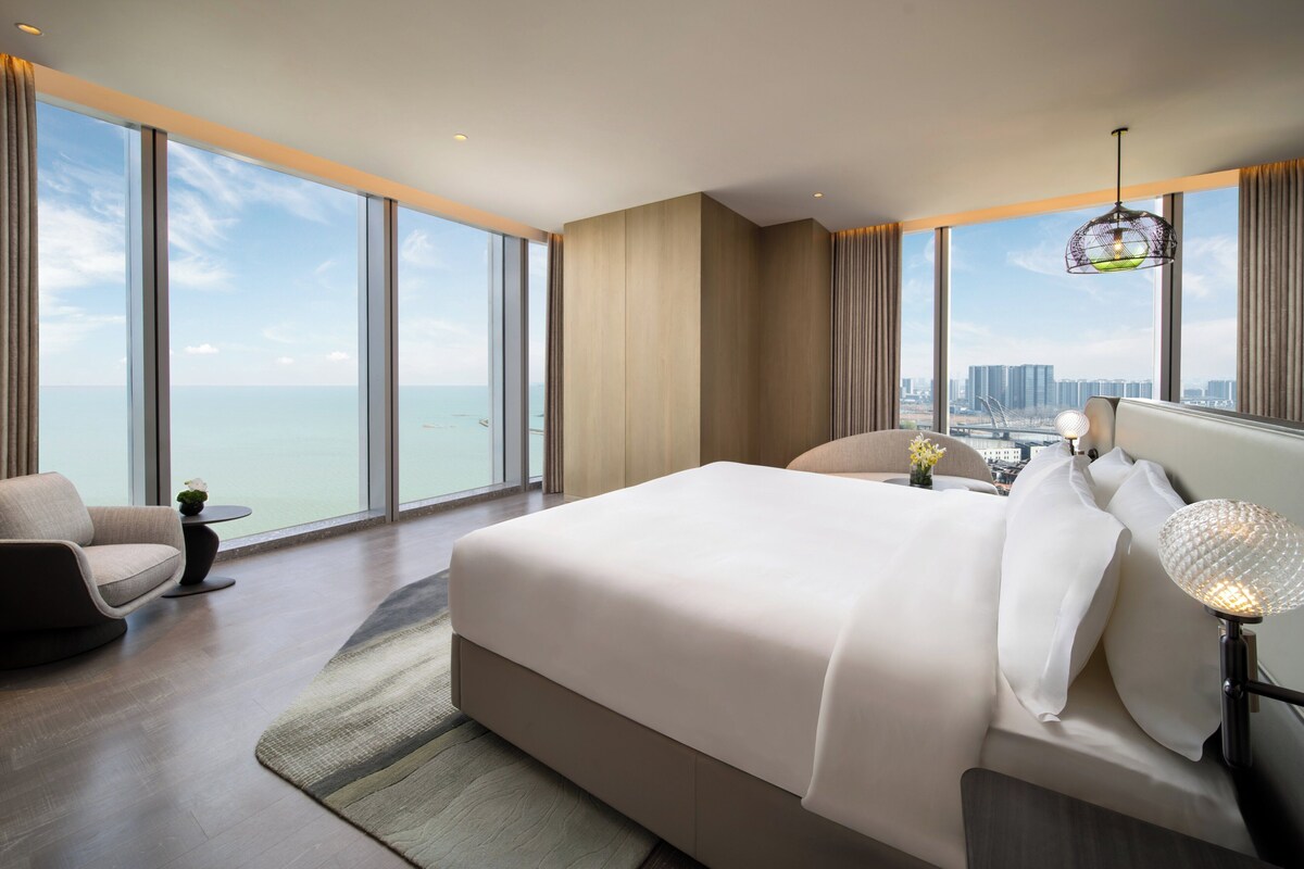 A modern hotel room with a large bed, a seating area, and a view of the city and sea.