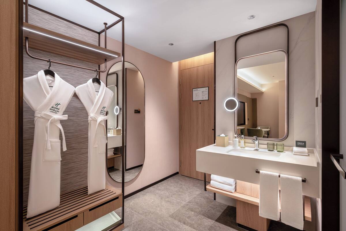 A modern bathroom with a wooden vanity, a large mirror, and two white robes hanging on a rack.