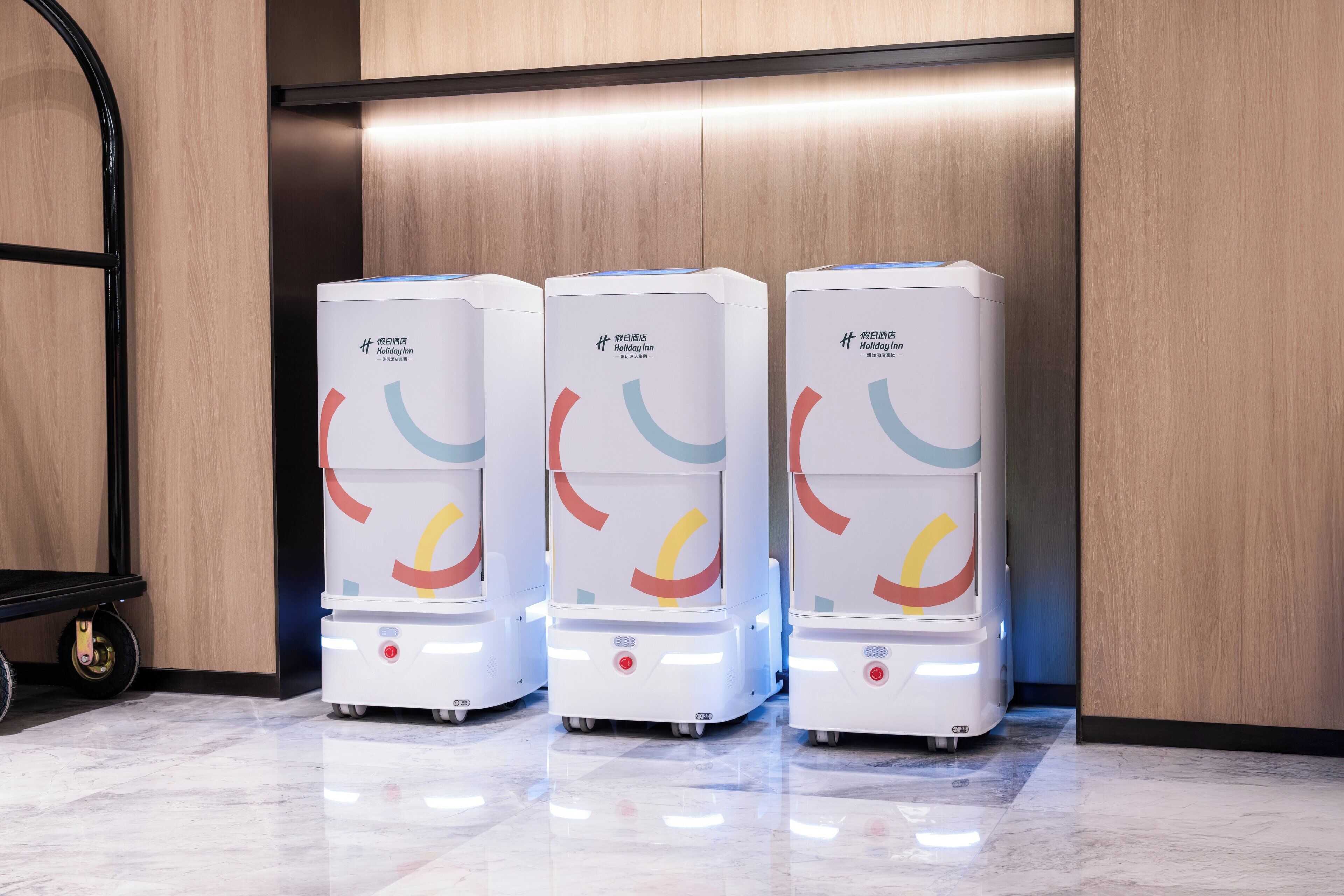 Three white self-service kiosks with colorful graphics, labeled "Holiday Inn," in a clean, modern lobby with wooden paneling and a marble floor.