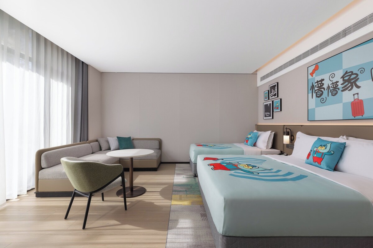Una habitación de hotel moderna con sofá, una pequeña mesa redonda y una cama con ropa de cama de temática de dibujos animados.