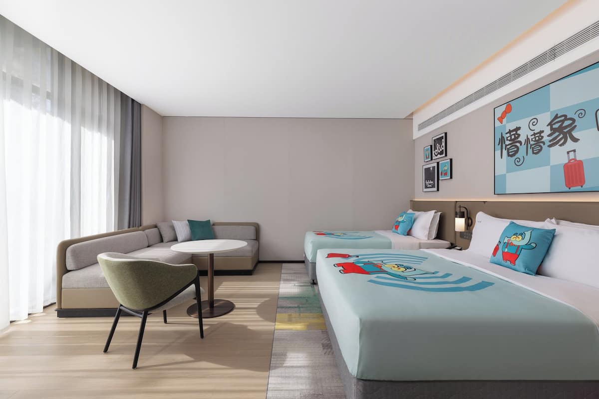 A modern hotel room with a sofa, a small round table, and a bed with cartoon-themed bedding.