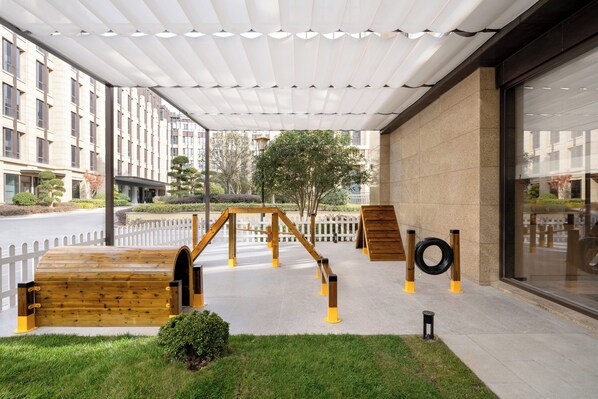 Un espace extérieur abrité doté d’une structure de jeu en bois, d’une clôture blanche et d’un bâtiment visible au fond.