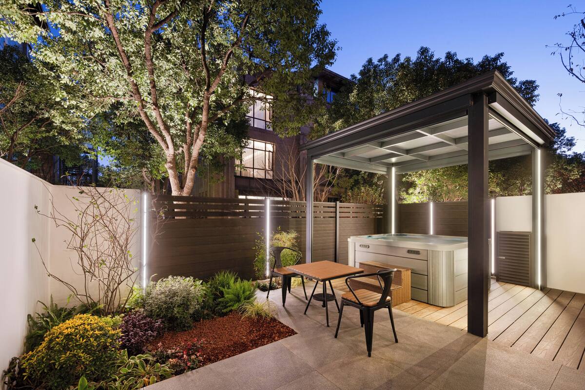 A patio with a hot tub, wooden table, and chairs, surrounded by a garden and a wooden fence.