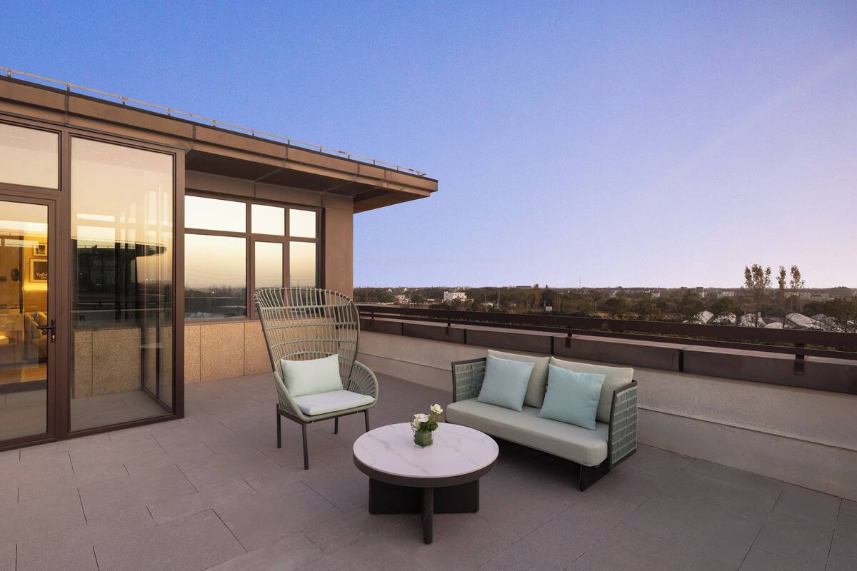 A rooftop terrace with a glass door leading to a room, a round table with a vase, and a light blue sofa with cushions.