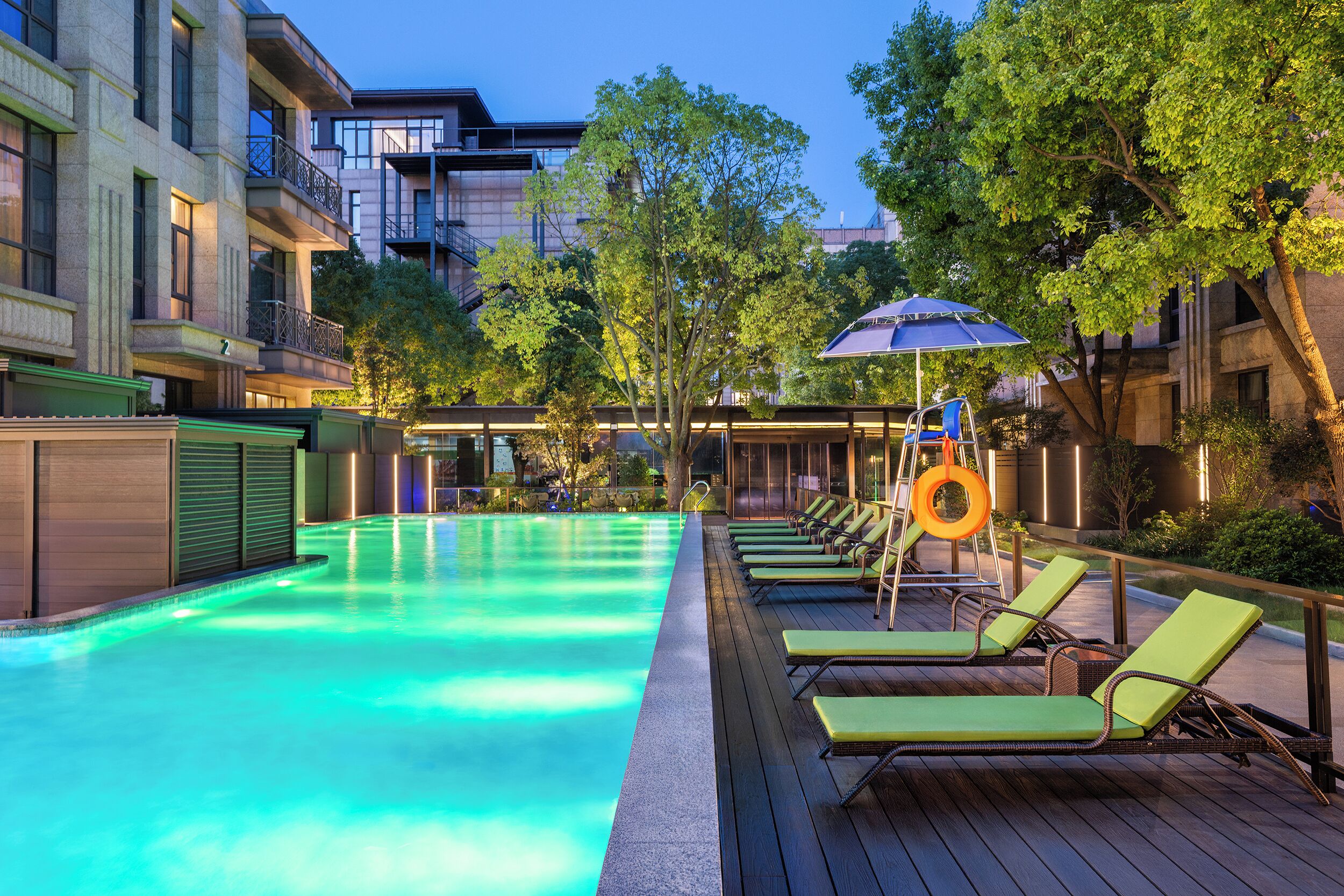 A swimming pool with lounge chairs and an umbrella, surrounded by trees and buildings.