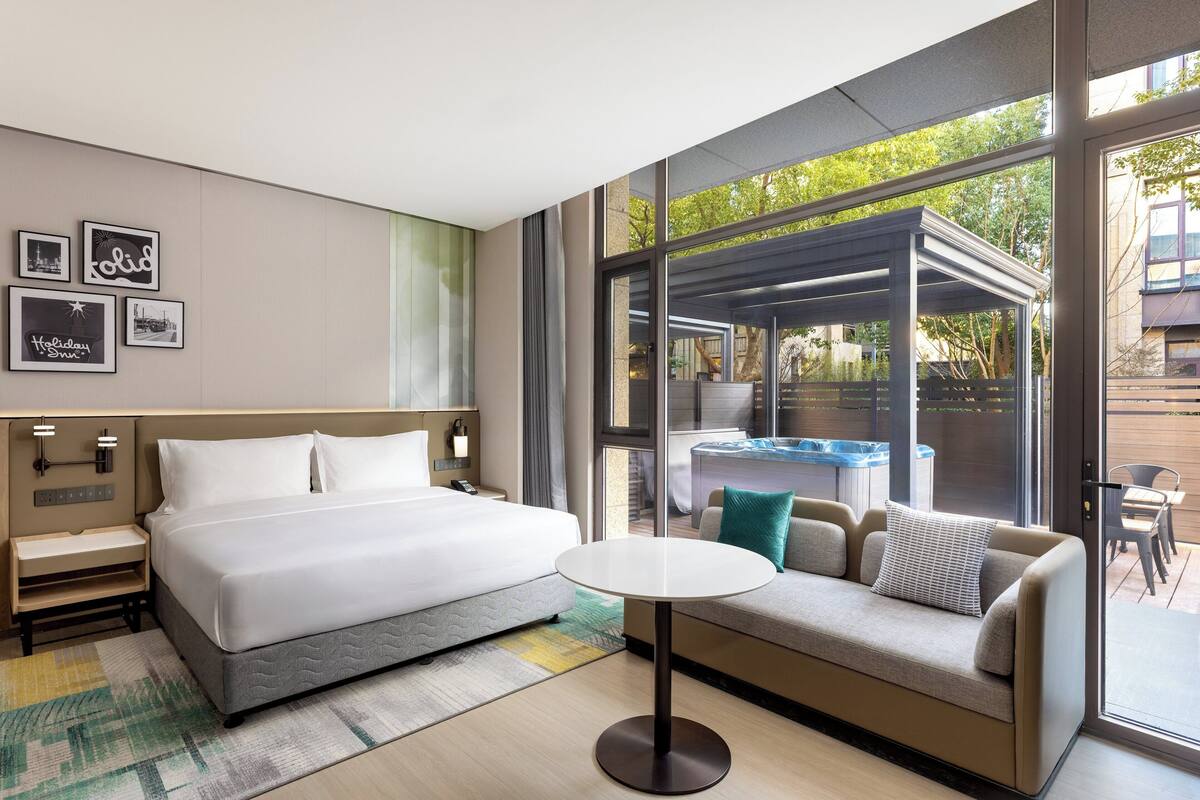 A modern hotel room with a large bed, a sofa, a small table, and a view of an outdoor hot tub.