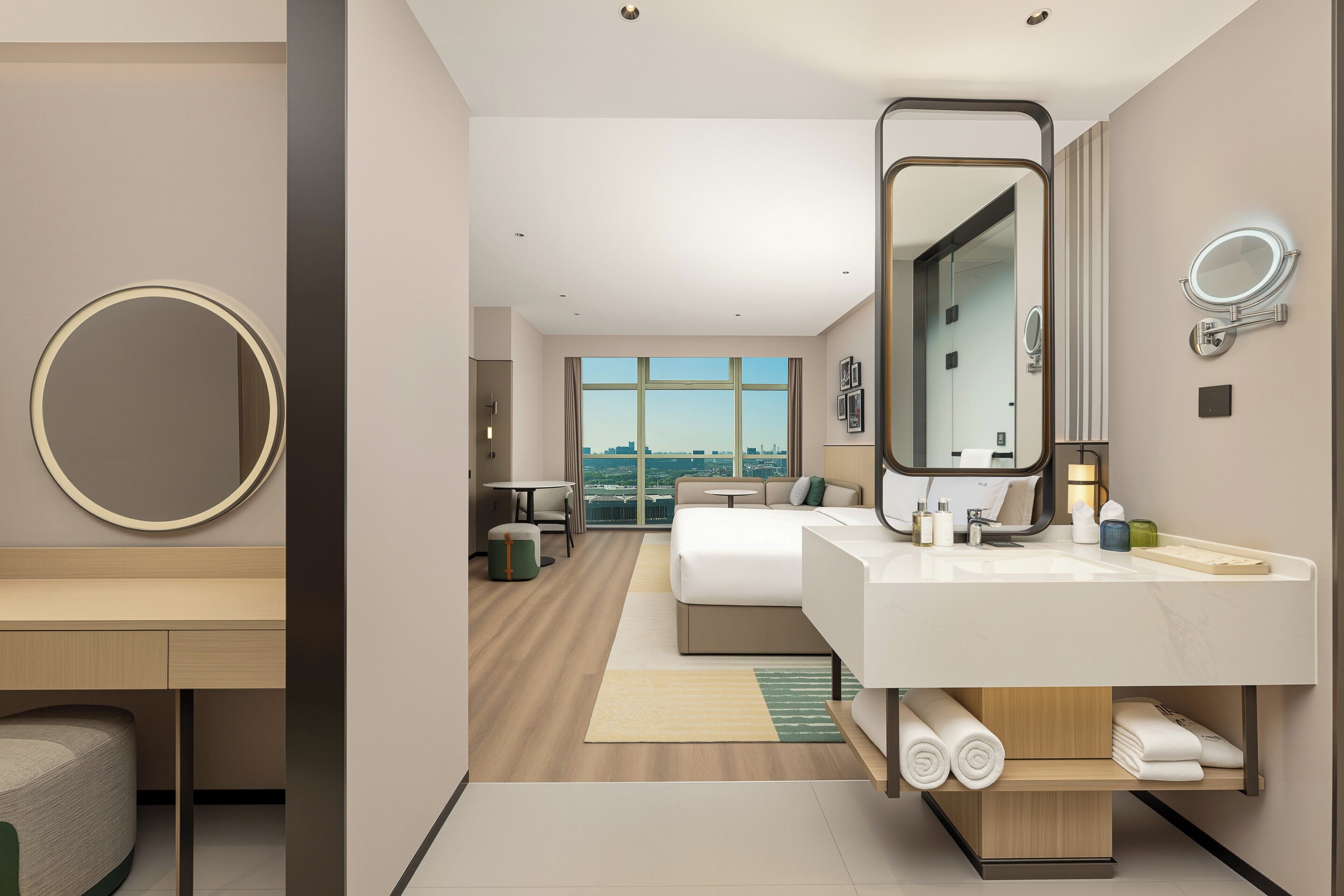 A modern hotel room with a large bed, a desk, a round mirror, and a bathroom with a large mirror and sink.