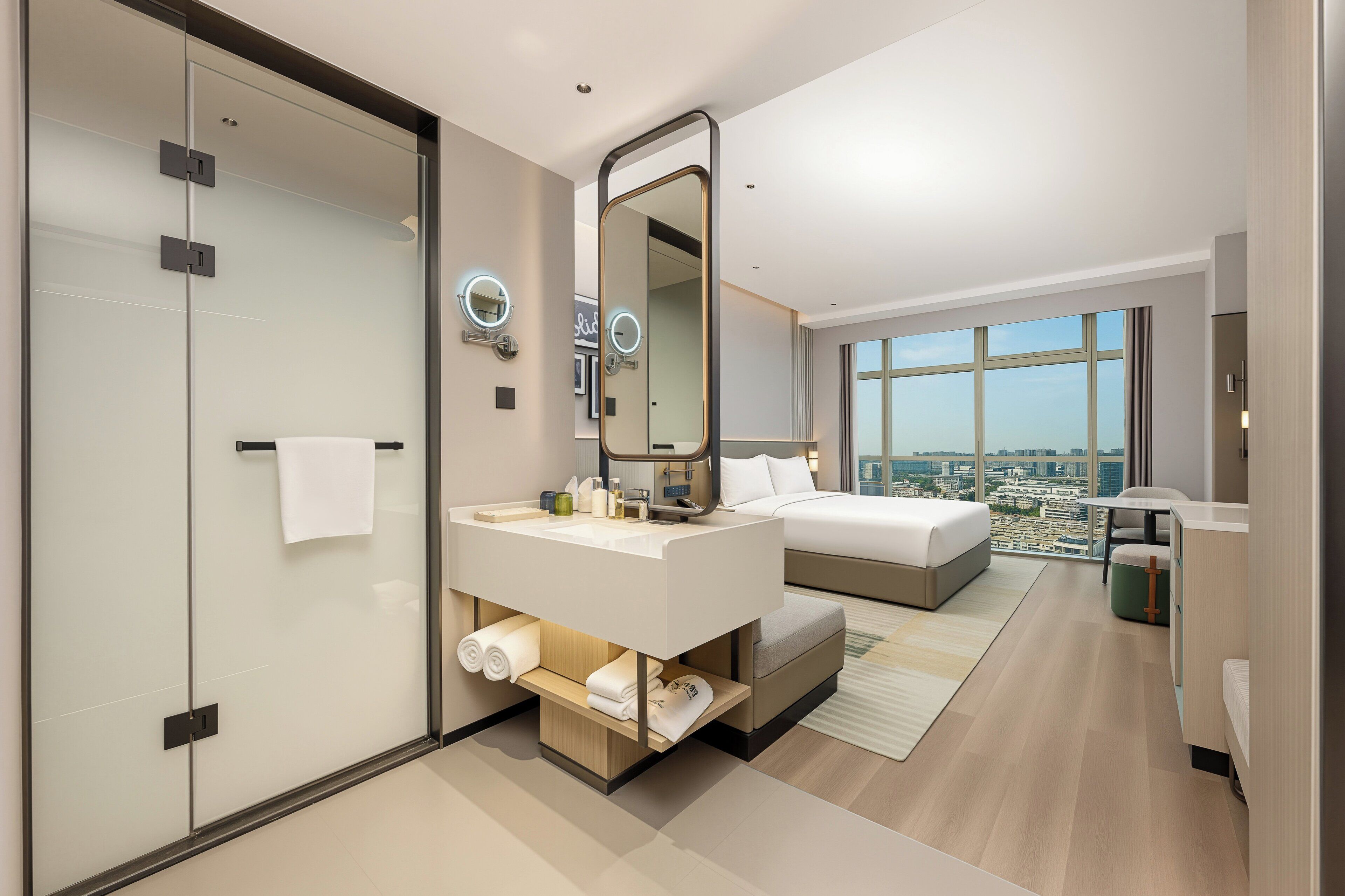 A modern hotel room with a large bed, a separate sitting area, a bathroom with a glass partition, and a city view.