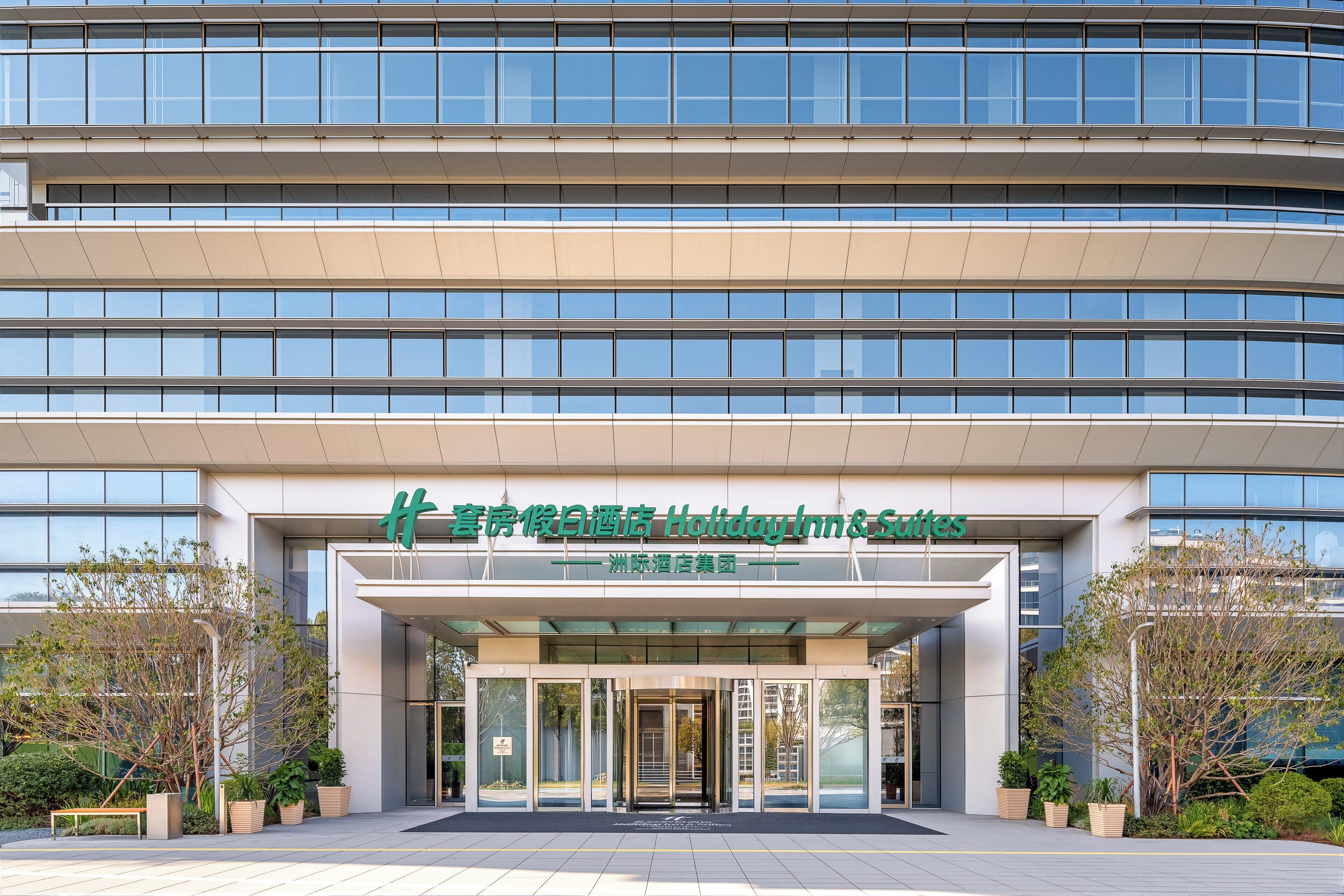 A modern hotel entrance with a glass facade, a sign indicating it's part of the Holiday Inn & Suites group, and a landscaped entrance area.